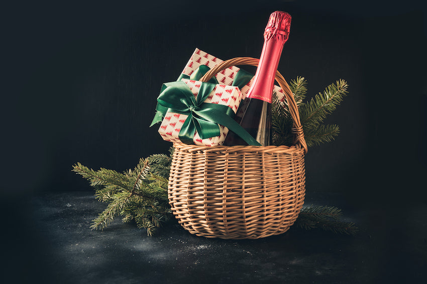 Una cesta de Navidad terminada, con variedad de dulces y bebidas, lista para regalar, con un lazo rojo y fondo festivo.
